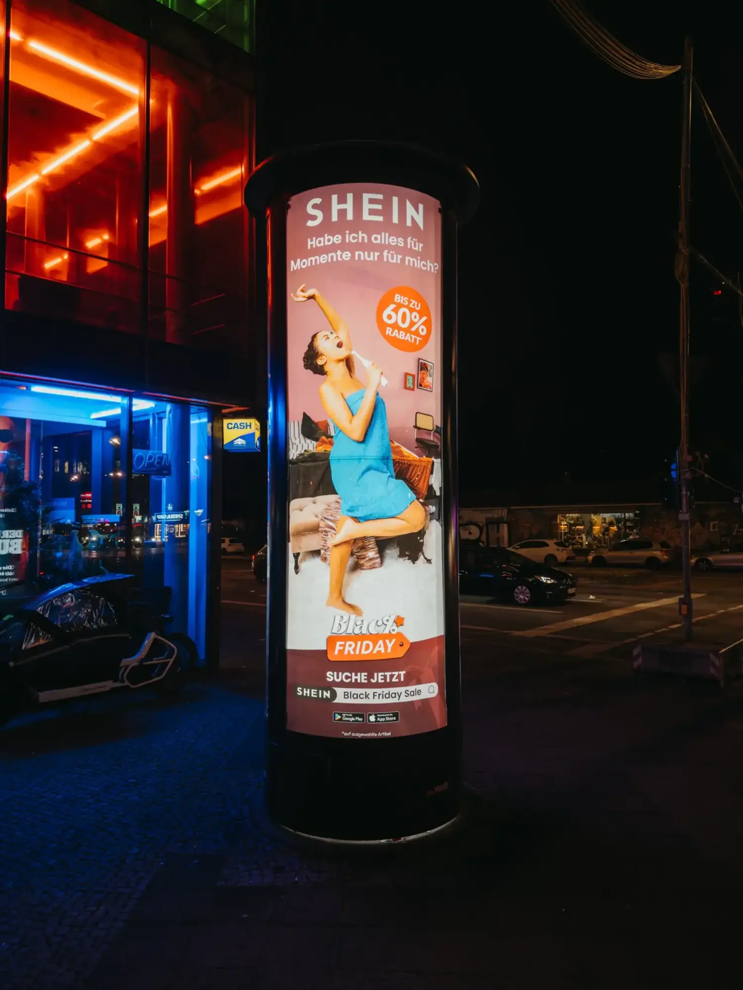 Campaign result: An illuminated advertising column in Berlin at night featuring the SHEIN Black Friday poster (Motif: Woman in towel).