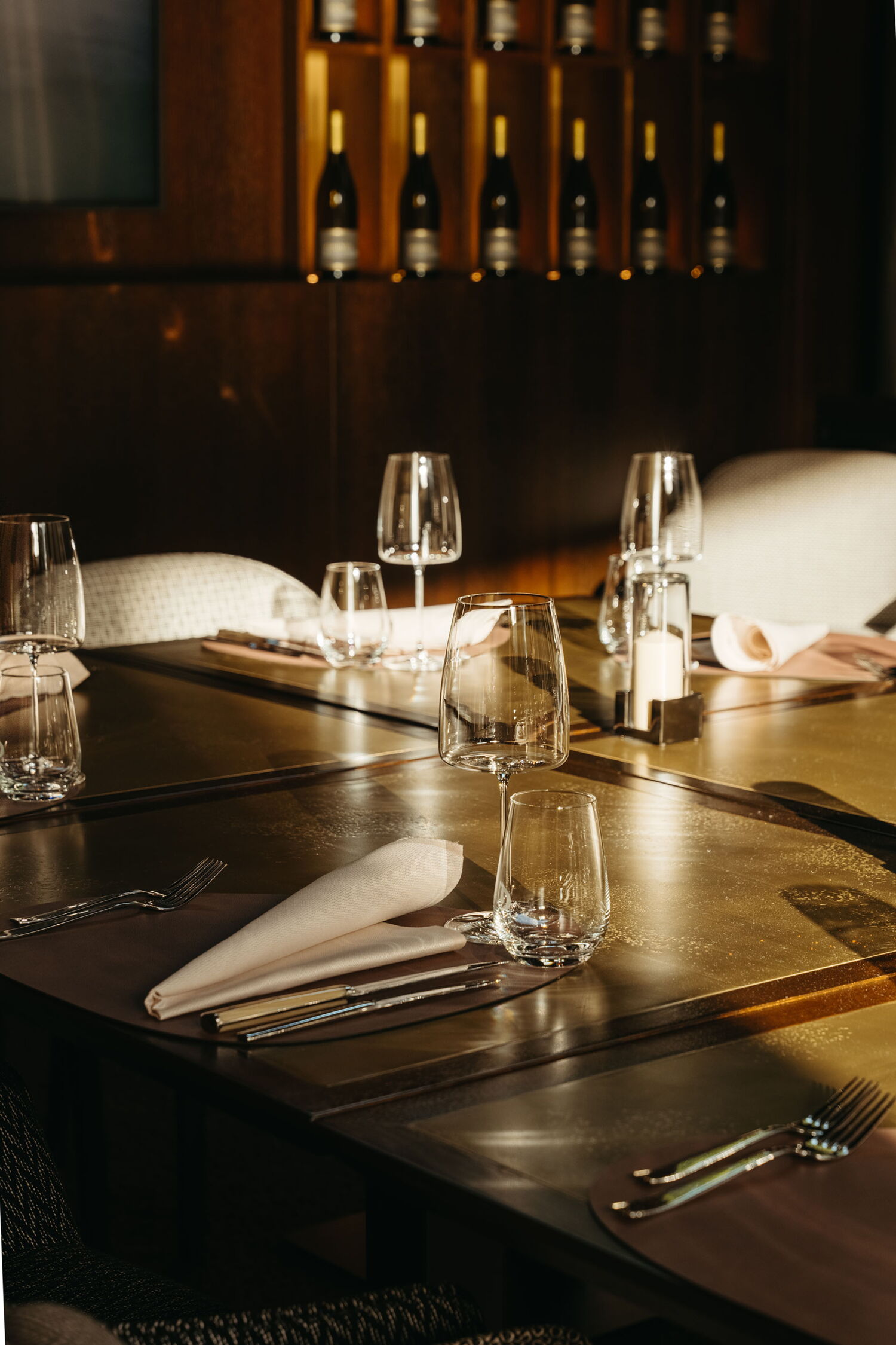 Close-up detail of an elegantly set table with wine glasses and cutlery, illuminated by warm, golden sunlight.