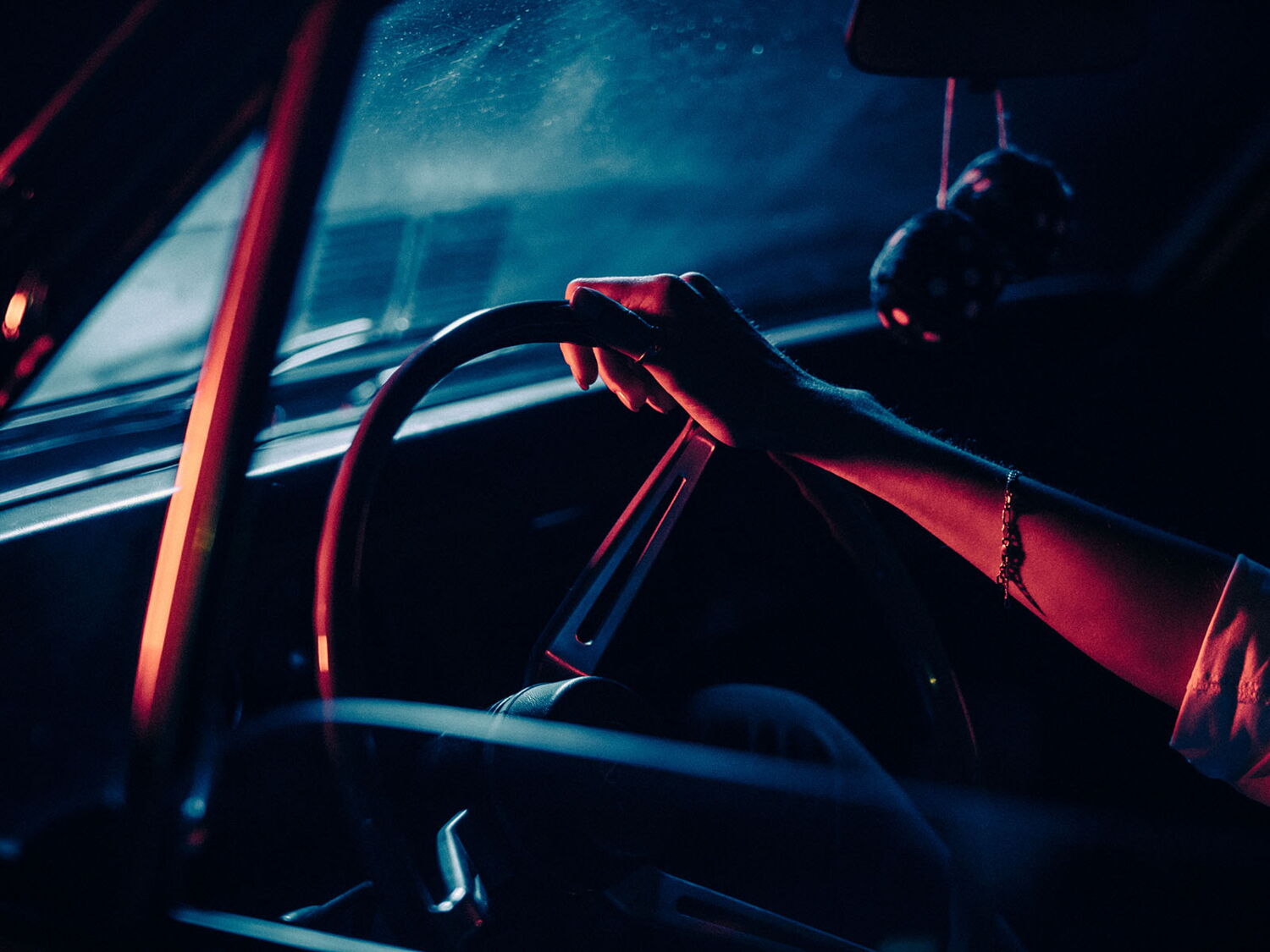 Cinematic close-up of hands on a car steering wheel at night, illuminated by a strong blue-red color contrast.