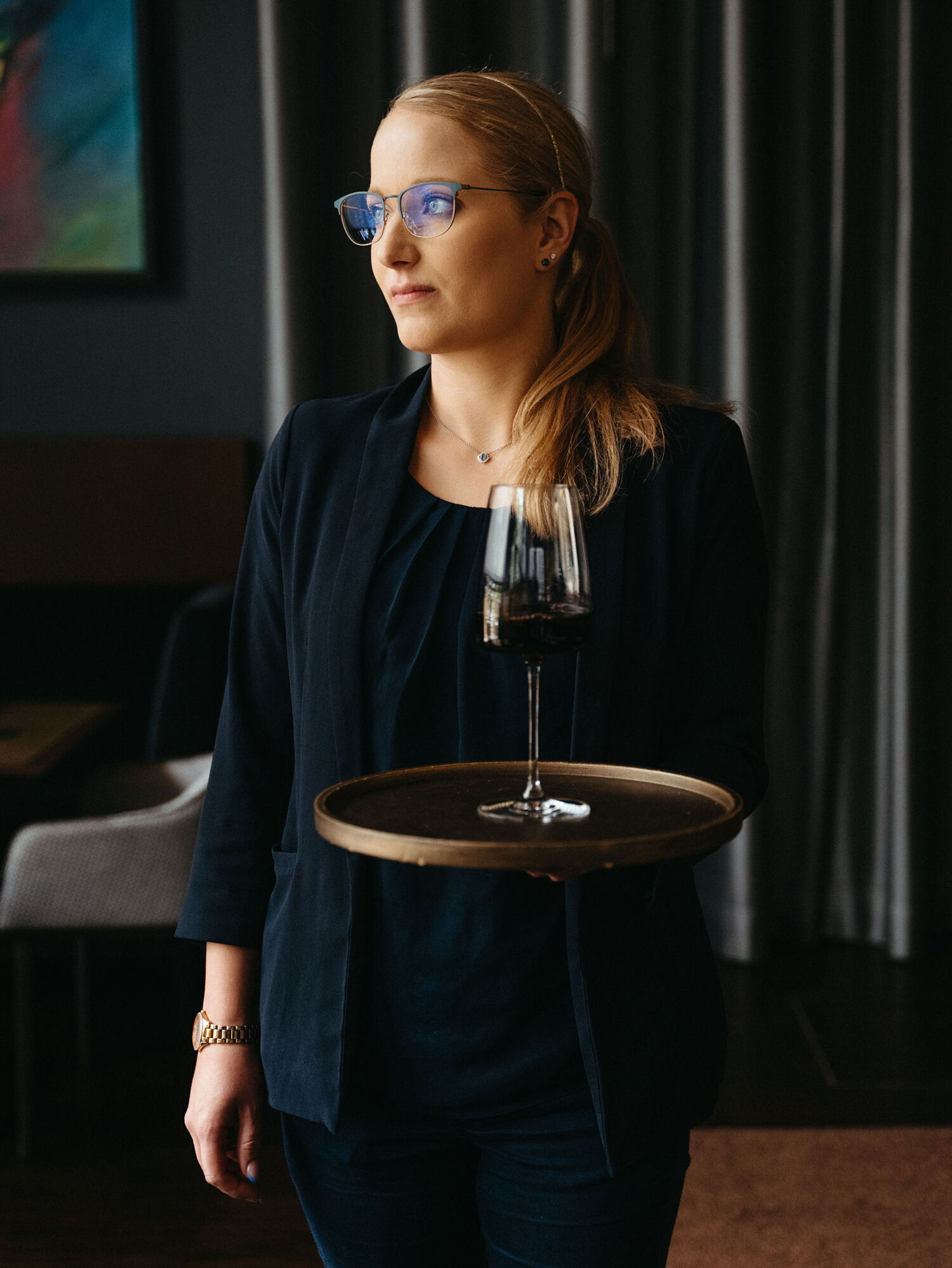 A service staff member with glasses elegantly holding a tray with a glass of red wine in the ambient light of the restaurant.