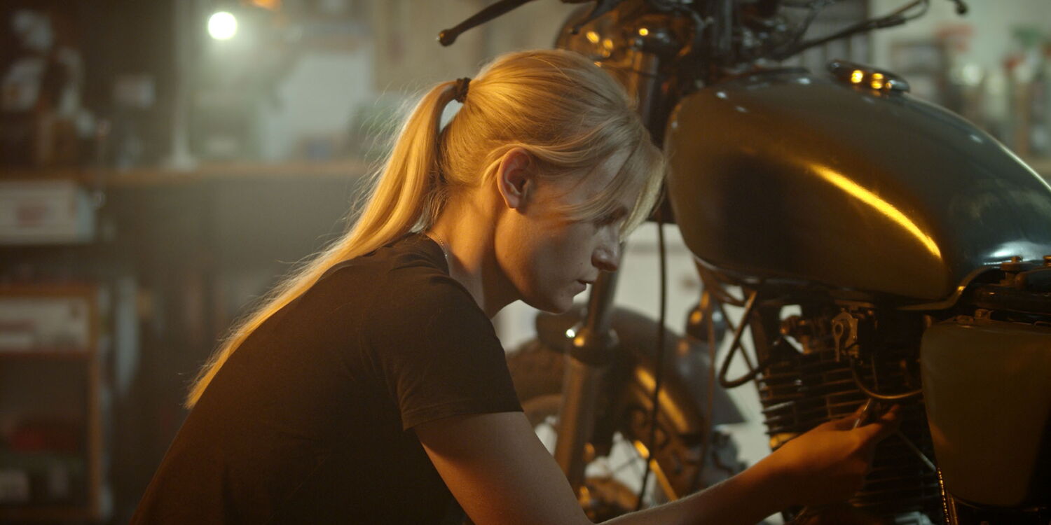 A woman crouching next to her classic motorcycle in a garage, inspecting the machine before a night ride.