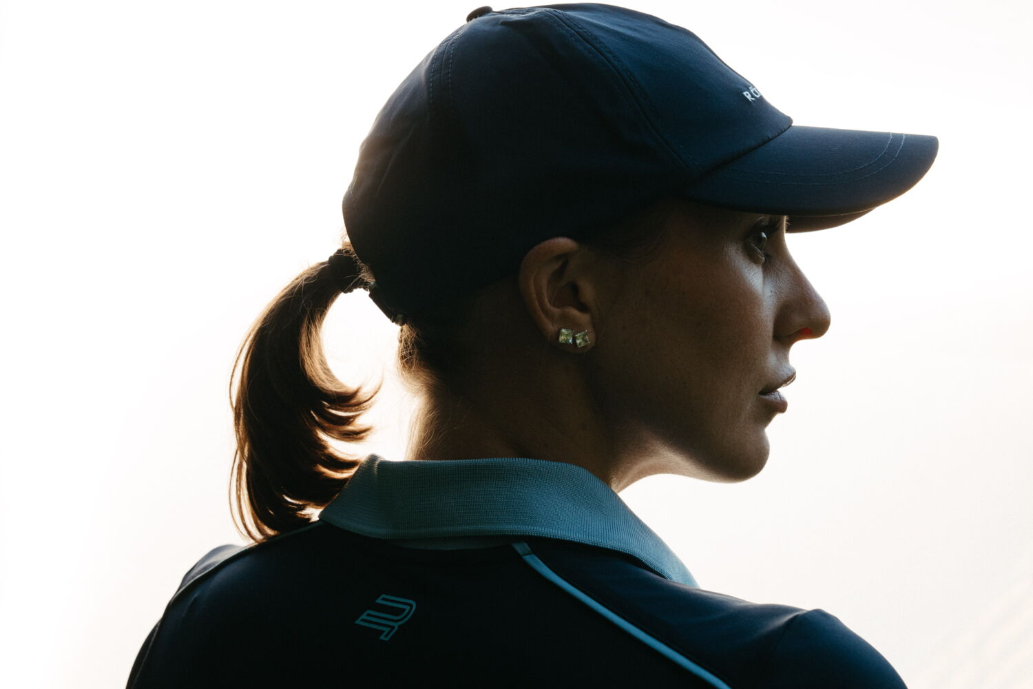 Profile shot of Laura Kowohl in strong backlight, highlighting the contours of her golf cap and earrings.