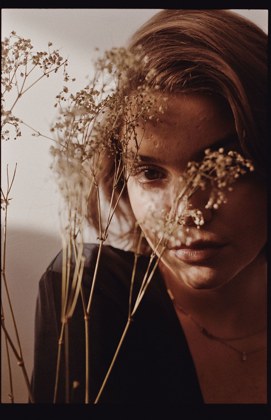 Gentle daylight portrait with an analog film aesthetic, photographed through blurred dried flowers in the foreground.