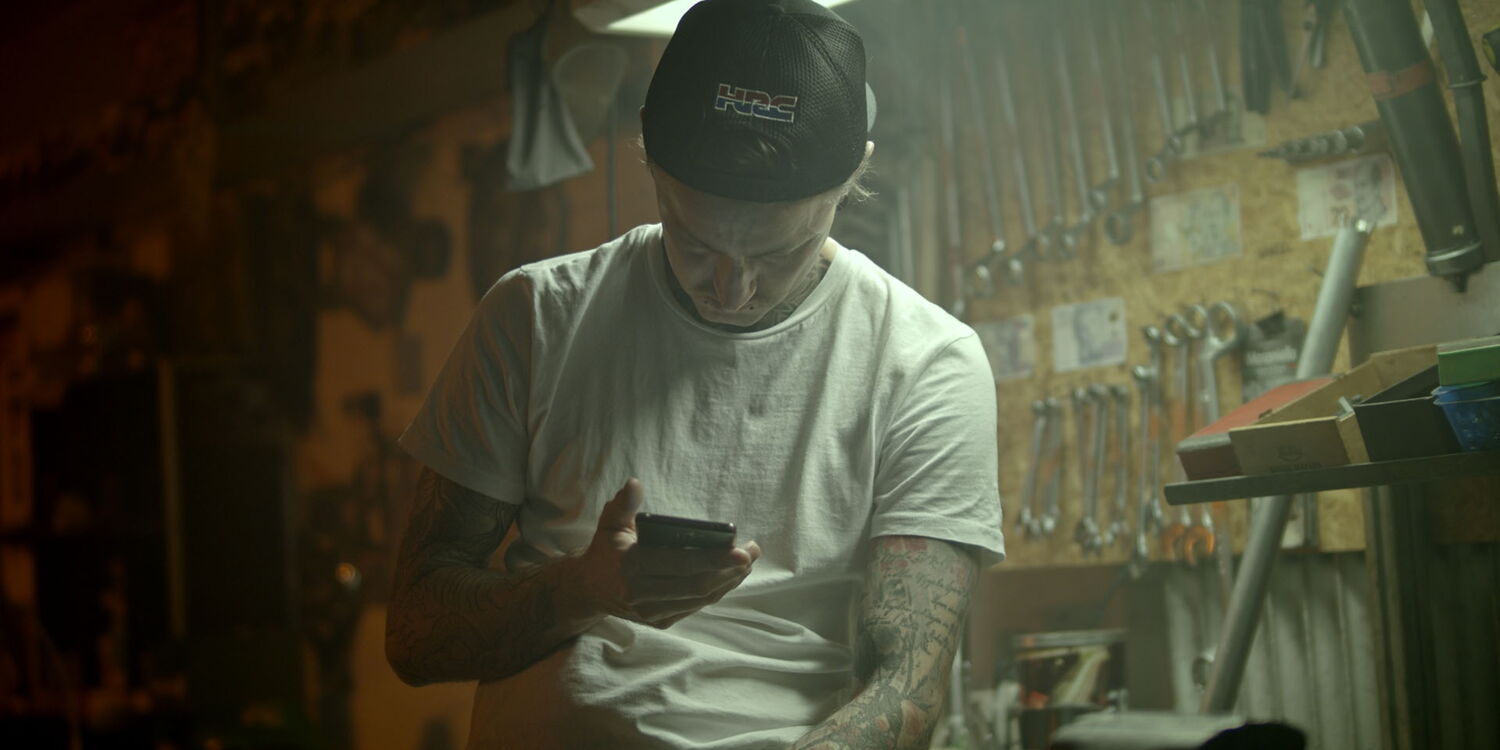 A tattooed mechanic wearing a cap looking at his smartphone, in front of a wall full of tools in an authentic workshop.