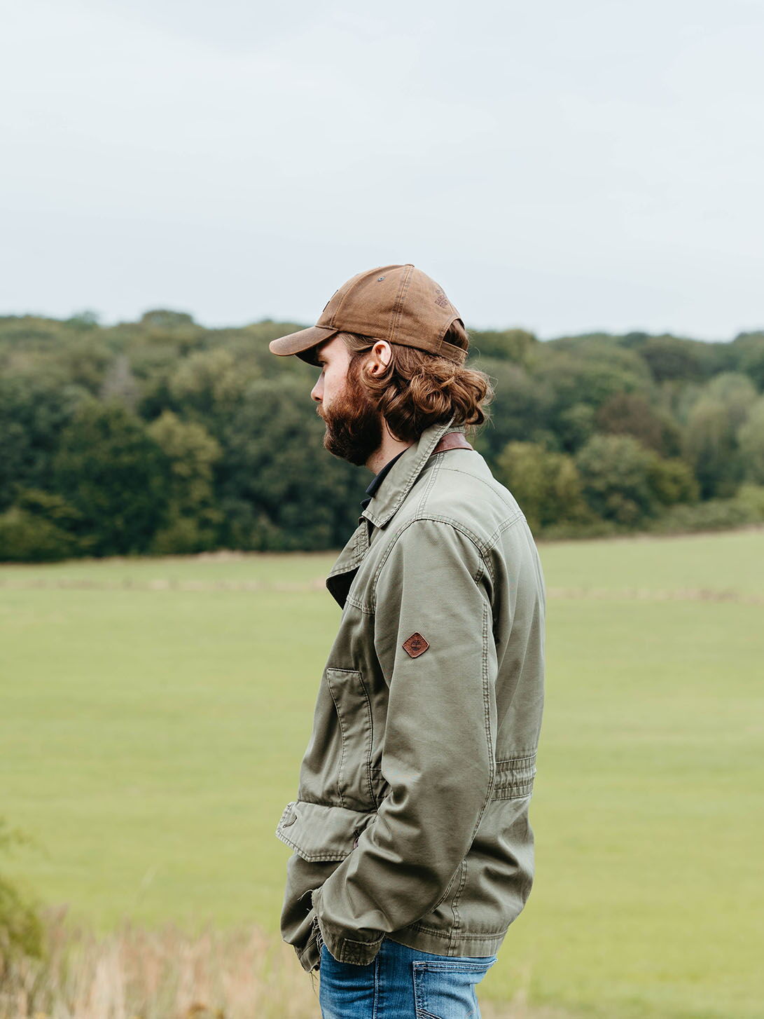 Profile of a young man with a cap and jacket looking over wide green fields.