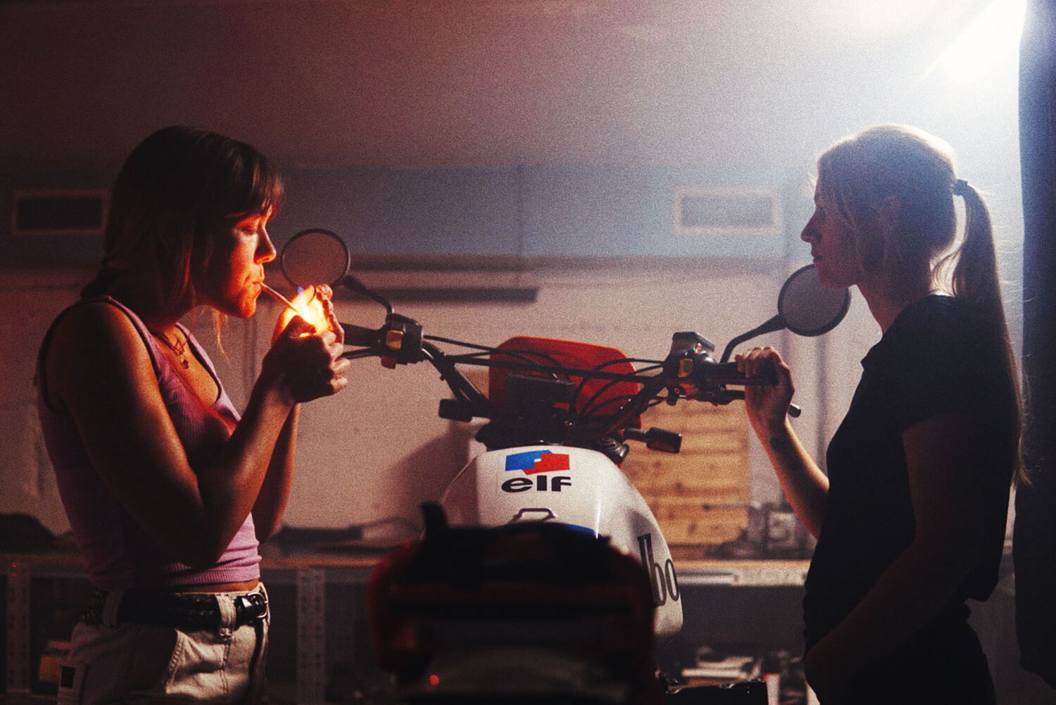 Two women sharing a cigarette in a dark motorcycle workshop, behind a bike featuring the Elf logo on the tank.