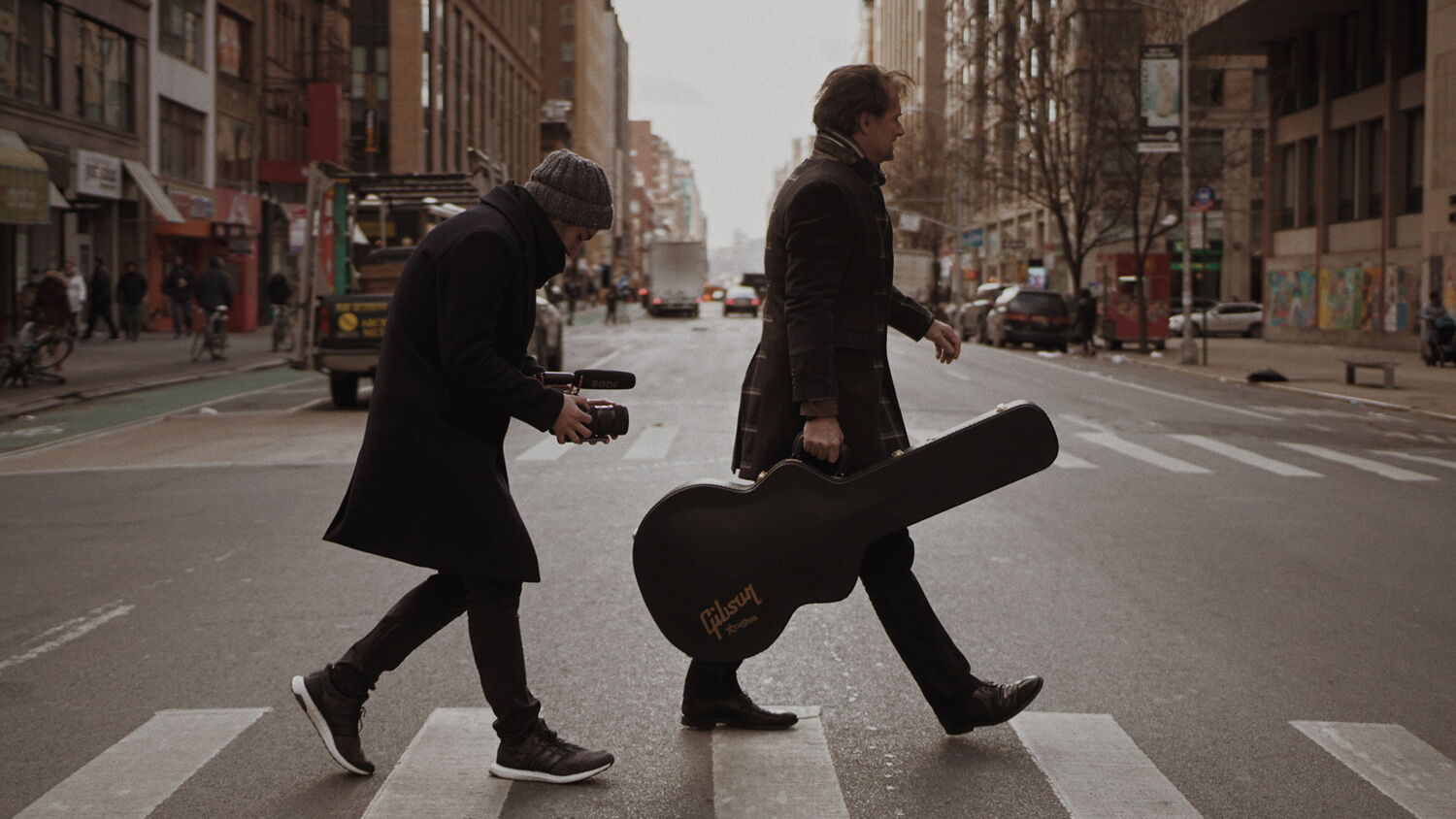 Jazz guitarist Oliver Doering crossing a street in New York City with his guitar case, followed by a cameraman.