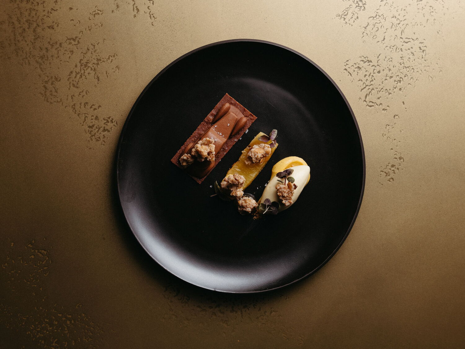Top-down view of a dessert trio on a black plate, consisting of a chocolate car, fruit element, and ice cream quenelle with crumble.