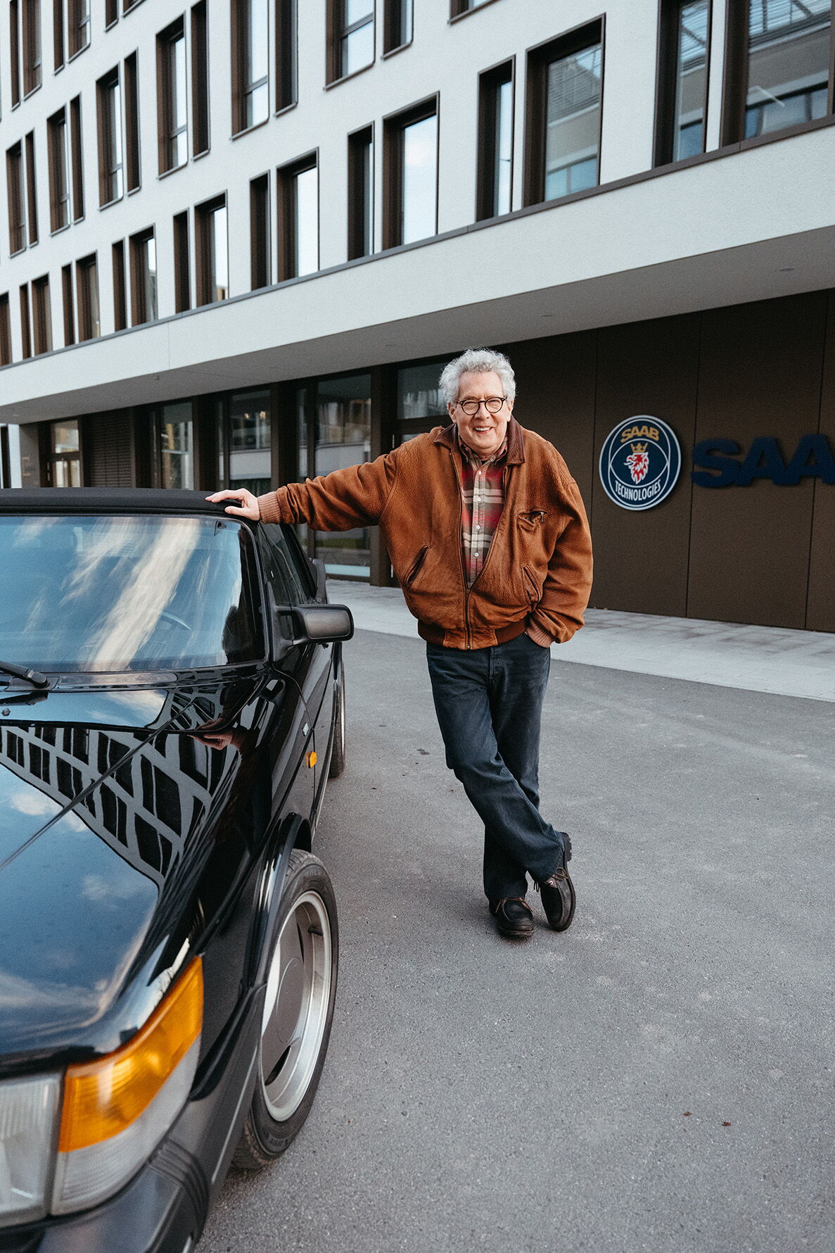 The protagonist leaning relaxed against his black Saab convertible in front of the modern Saab company building.