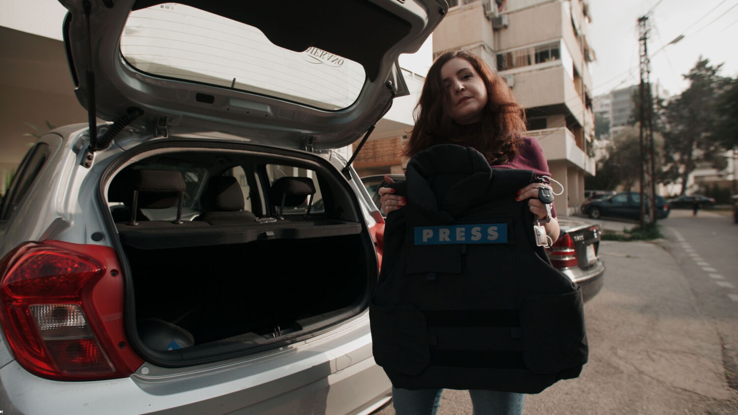 A protagonist holding up her bulletproof press vest from a car trunk, symbolizing the dangers of reporting in conflict zones.