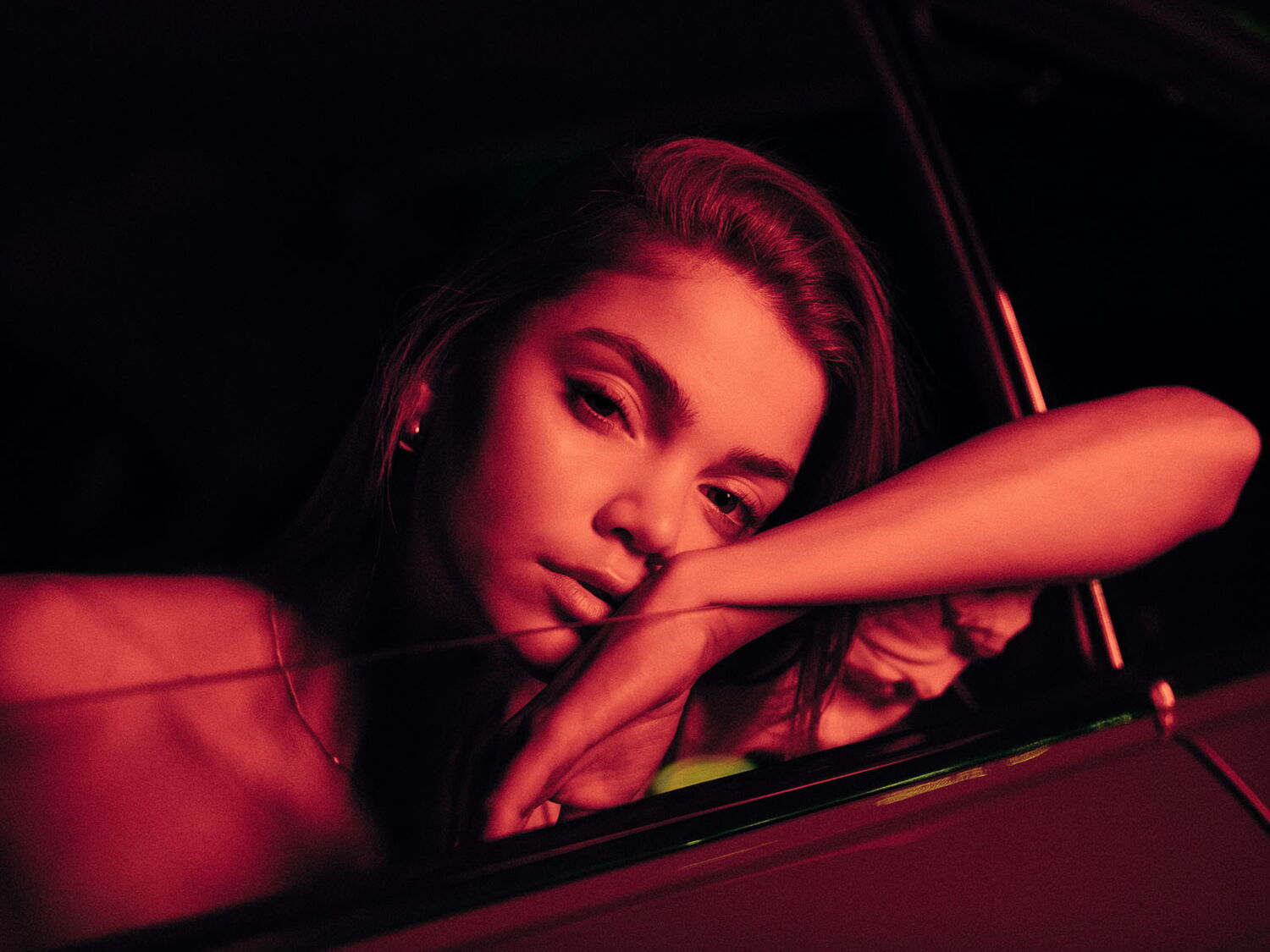 A young woman dreamily resting her head on a car window frame, bathed in atmospheric red ambient light.