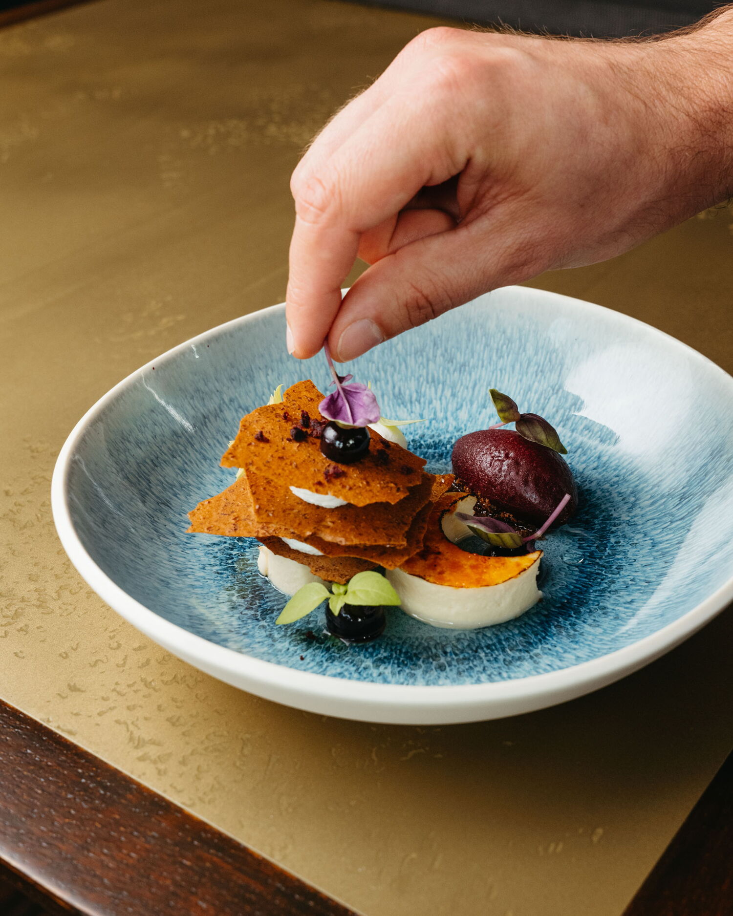A chef's hand carefully placing a purple leaf as a garnish on a complex dessert featuring sorbet and crispy layers.