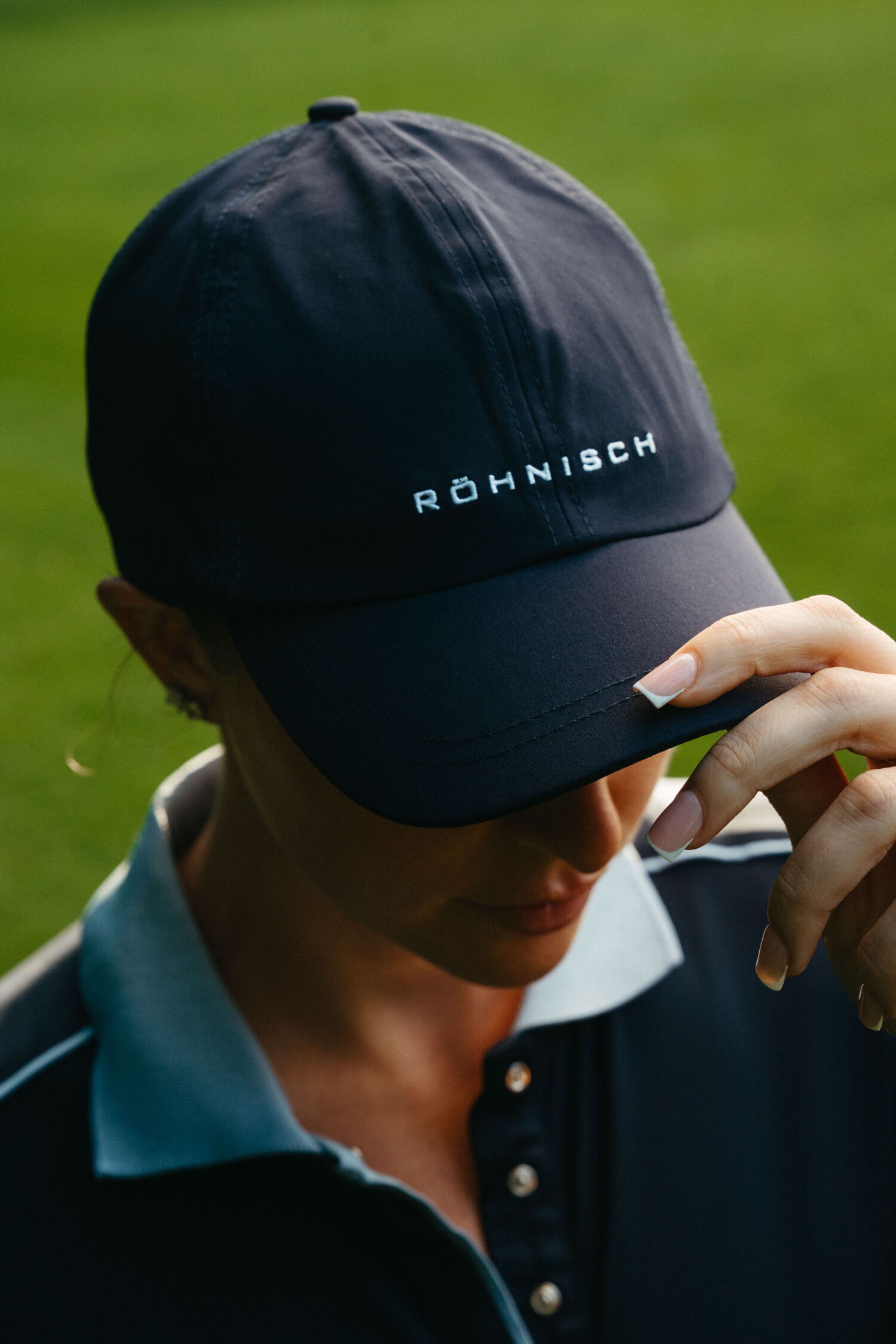 Close-up detail of a hand touching the brim of a navy blue golf cap featuring the embroidered Röhnisch brand logo.