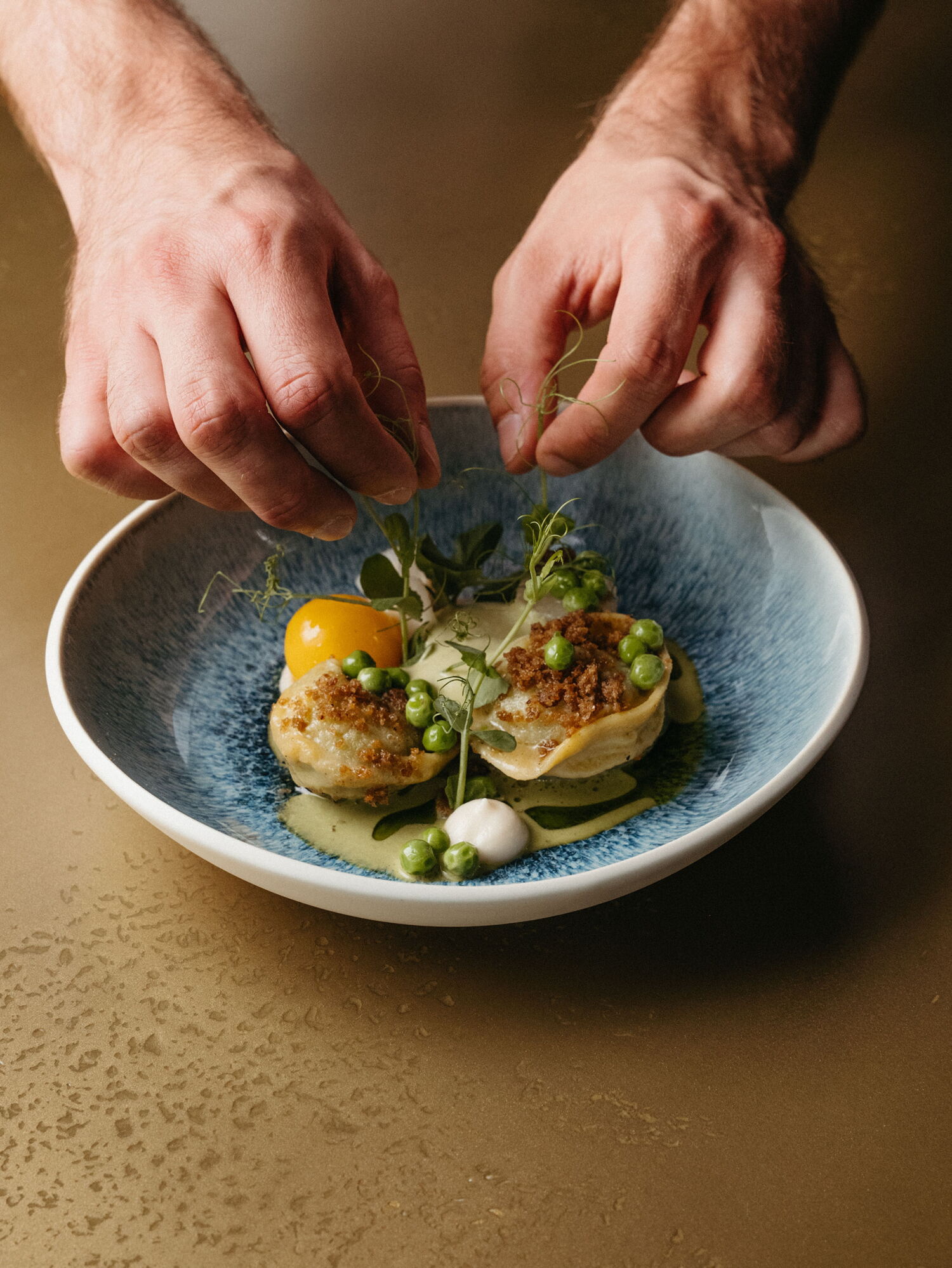 Close-up of a chef's hands precisely placing fresh herbs on a gourmet dish featuring peas and foam.