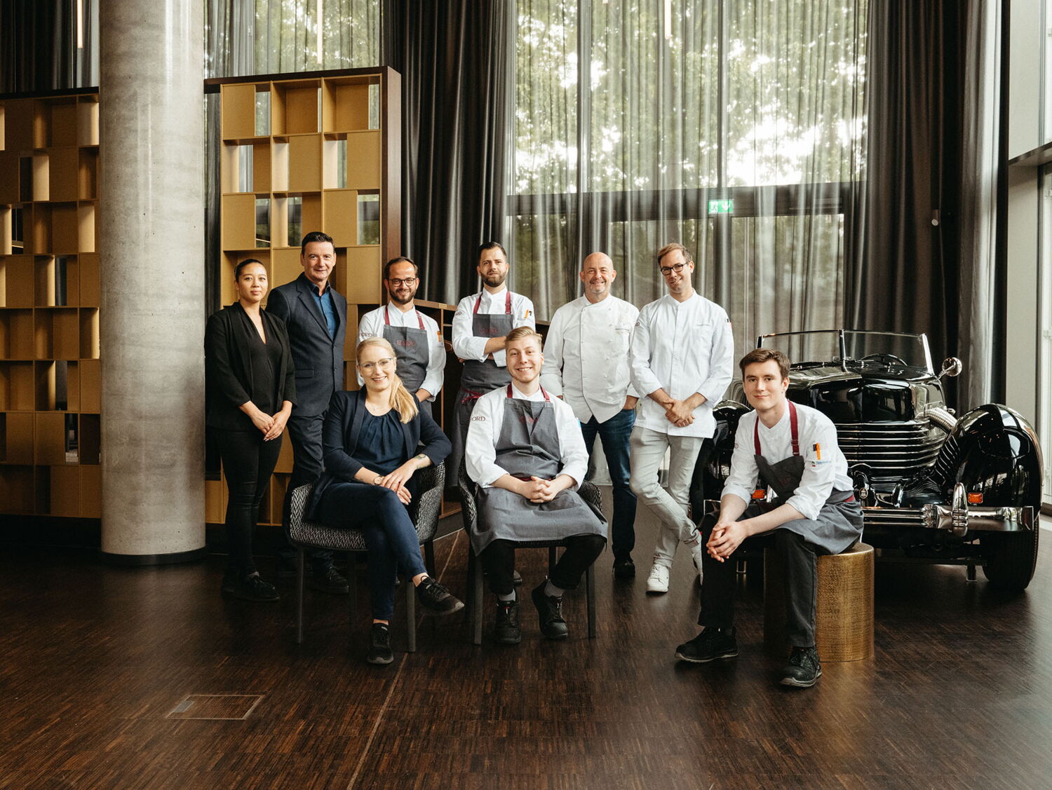 The kitchen and service team of The Cord restaurant posing together in front of a vintage car inside the glass-walled dining area at EUREF-Campus.