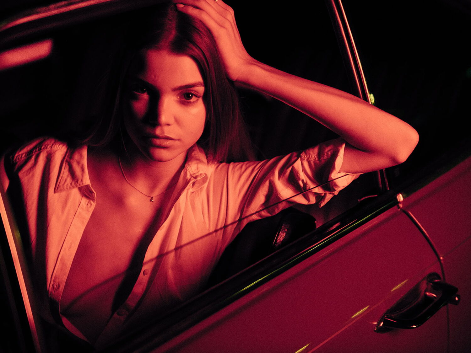 Editorial portrait of a young woman leaning out of a car window at night, illuminated by dramatic red neon light.