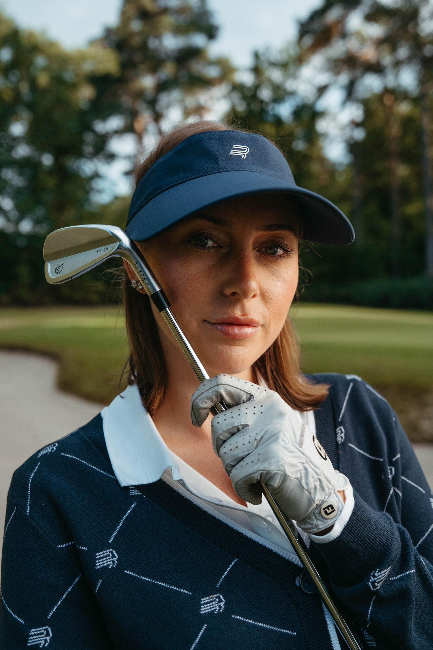 Portrait of Laura Kowohl holding an iron club close to her face, wearing a blue Röhnisch visor and golf glove.