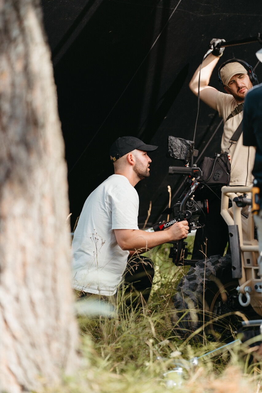 Camera operator filming from a low angle in tall grass to capture the military vehicle dynamically.