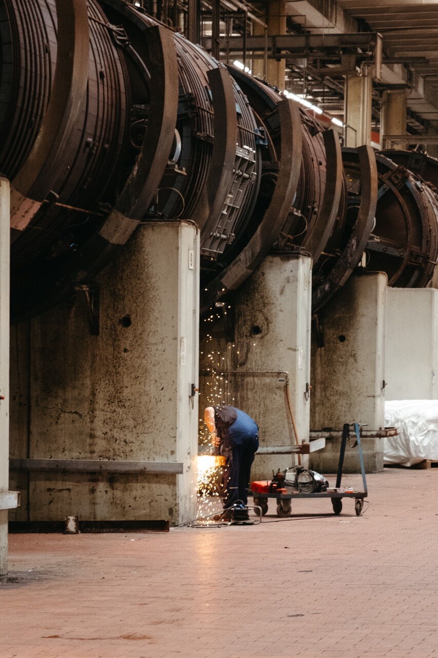 A worker metalworking with flying sparks in front of giant industrial drums in a factory hall.