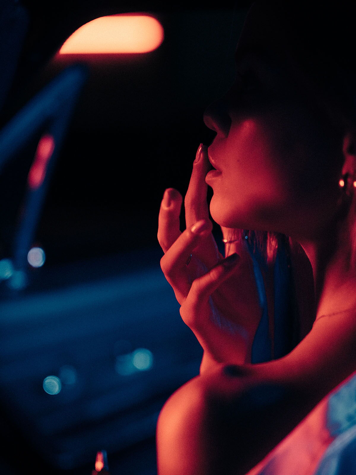 Side profile of a young woman in the passenger seat at night, accented by blue street light and red reflections.