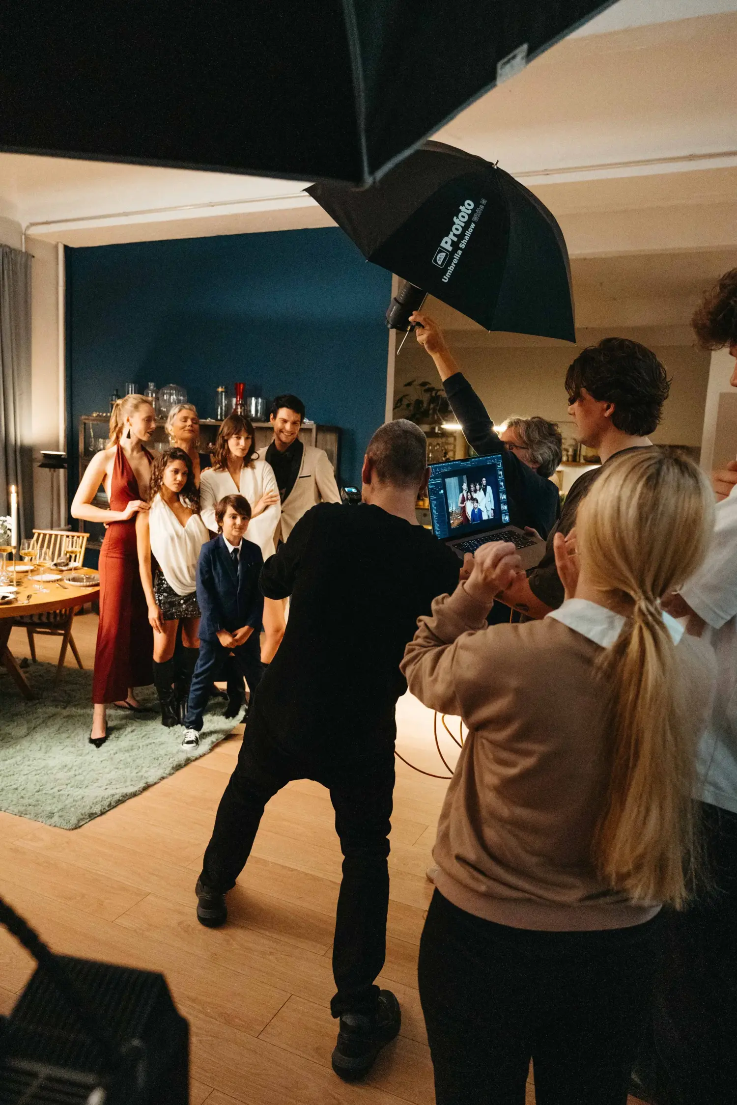 Production crew on set: Digital Operator checking the shot on a laptop, with the cast posing for a group photo in the background. Lighting with Profoto umbrellas.