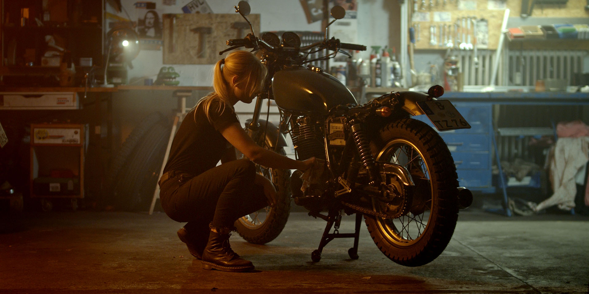 Eine Frau arbeitet konzentriert am Motor eines Custom-Bikes in einer Garage, beleuchtet durch stimmungsvolles Gegenlicht.